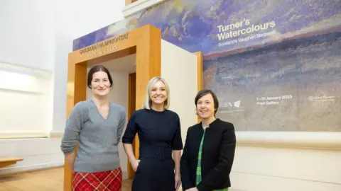 Three women stand in front of the entrance to an art gallery. They are smiling and two are wearing tartan. There is a Turner watercolour behind them, advertising the exhibition