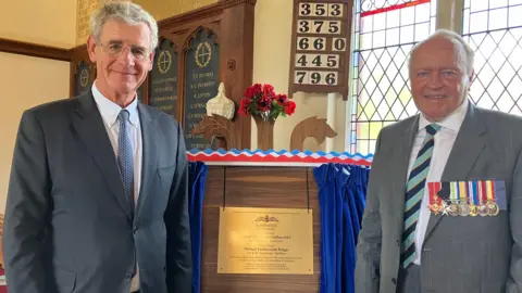 Two men with grey hair wearing suits, one of which has a row of military medals on, standing next to a gold plague about the size of an A4 piece of paper. They're in a chapel and a stained glass window can be seen behind them.