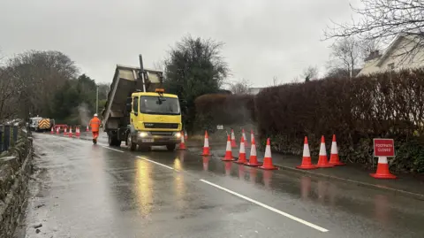 A yellow dumper truck on a wet road, with traffic cones marking out a section of roadworks. The road is bordered by a tall, browned hedge which borders a housing estate. There is a row of traffic cones in front of it.