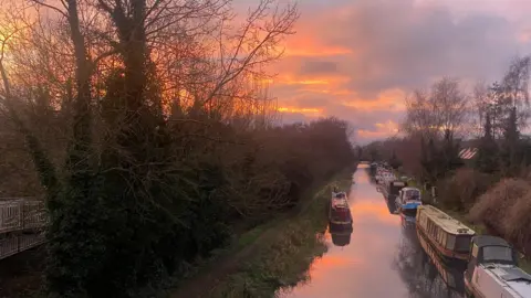 Carla A canal runs through the picture with several barges moored up on the right-hand side. There is a footpath and a lot of trees on the left-hand side of the canal. Overhead, the sky is a golden yellow colour as the sun sets.