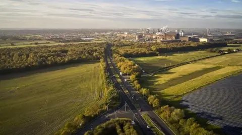 North Lincolnshire Council Aerial view of Mortal Ash Hill with trees along the side of the road and the town's steelworks in the distance.