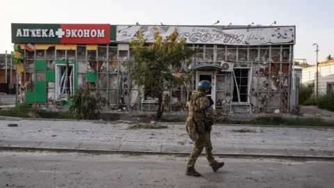 Getty Images A soldier in fatigues and a helmet walks past a dilapidated building