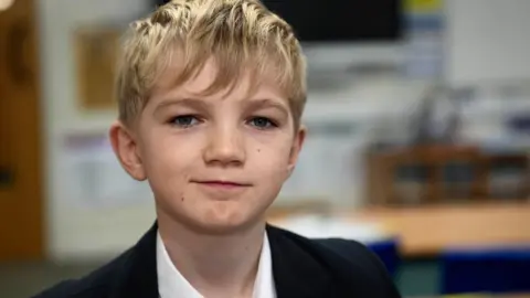 John Fairhall/BBC Miles, a Year 8 pupil who has short fair hair brushed over his forehead. He is wearing  a dark jacket and a white shirt. He is sitting in a classroom, blurred out behind him