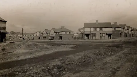 Wolverhampton City Archives A black and white picture showing houses which are two-storey \and are mainly semi-detached. In the front of the photo is a large area of ground which is empty. 