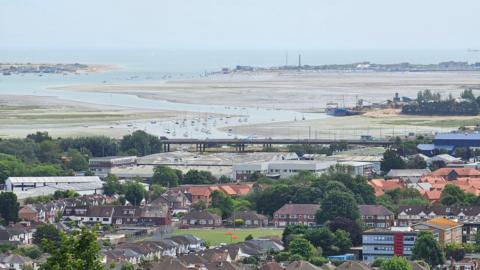 FRIDAY - A view from above over Langstone Harbour in Portsmouth. In the foreground are the roofs of homes and several industrial units. Behind cars drive over a causeway over the top of the harbour and behind the harbour stretchers away. There are mud flats at low tide with two channels of water running out into the Solent. Several boats are moored up and in the distance you can see the entrance to the harbour and Hayling Island.