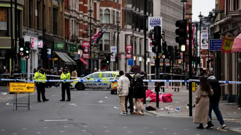 PA Media Members of the public walk past a police cordon on Shaftesbury Avenue. Two police officers are standing behind the cordon. There is a yellow sign that says 'diverted traffic' and an arrow pointing left which has been placed in front of the cordon.