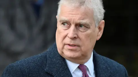 A man with white hair looks off to the side of the camera. He is wearing a dark blue jacked, a white shirt and a crimson-coloured tie.