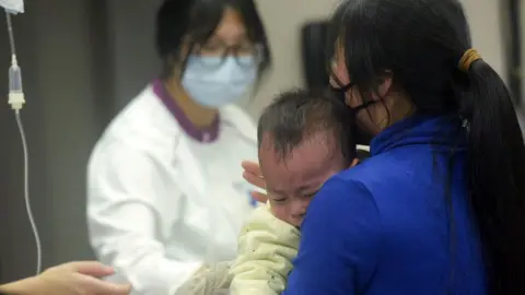 A baby wearing a yellow babygro is held by his masked mother as he receives care at the paediatric department of a hospital in Hangzhou, eastern China's Zhejiang province, on 6 January 2025.