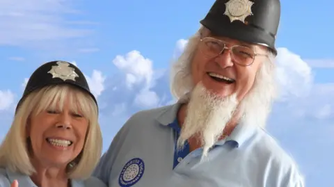 Clive St James Clive, with white beard, stood with his wife, Jackie, both wearing blue shirts in front of a cloudy blue sky. They are both wearing traditional style police helmets. 