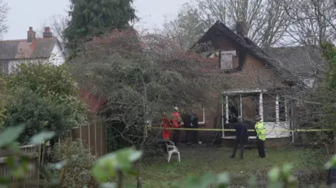 A back garden and house, with fire officers and a police officer standing close to the building, which has yellow police tape around it. The roof and bricks on the first floor are blackened