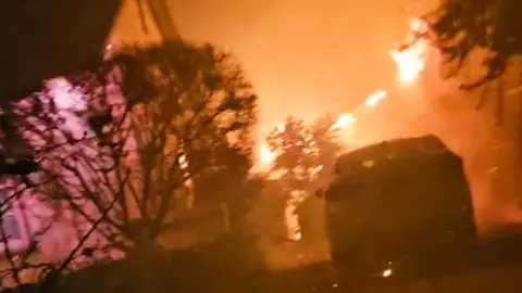 A blazing fire is seen engulfing a garage with a car parked in front of it