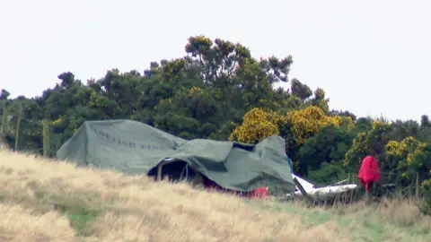 A green tarpaulin covers the wreckage of a small plane. Some white metallic parts protrude from under the cover. There are trees and bushes behind and dry white grass in the foreground.