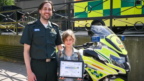 Emergency call handler Matthew Hawkins with Farah Alim-Palmer, who holds a certificate. They stand in front of a London Ambulance motorcycle at the emergency service's headquarters in Waterloo.