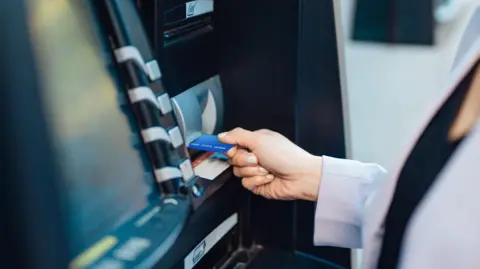 A woman getting money out of a cash machine, she is holding a bank card, and is putting it into the machine. 
She is wearing a light jacket, only her arm and part of her body is in view. 