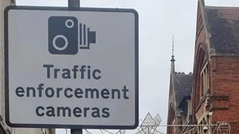 A "traffic enforcement cameras" sign. It features a picture of a traditional camera in black on a white background. A red and black building is to the left and a blue bus is to the right.