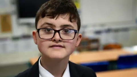 John Fairhall/BBC Harry a Year 8 pupil who has short dark hair brushed over his forehead. He is wearing dark framed glasses, a dark jacket and a white shirt. He is sitting in a classroom, blurred out behind him