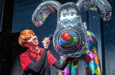 A woman polished a large Gromit statue at the launch of the Gromit Unleashed 3 trail in Bristol. The statue has a multicoloured sparkly pattern. The woman is wearing a red and white shirt and a black dress over the top and she has red hair cut in a bob style