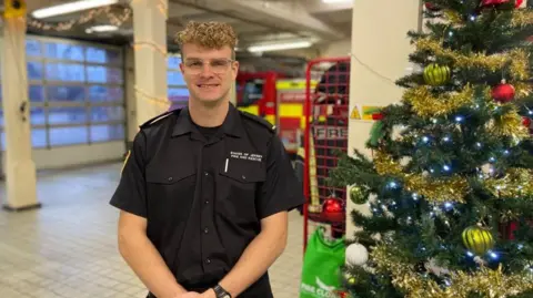 Firefighter Michael Milner stands next to a decorated Christmas tree, which is on the right hand side, in St Helier Fire Station. He is wearing a black short sleeve shirt and has glasses on. He has short curly blonde hair. A fire engine is in the background.