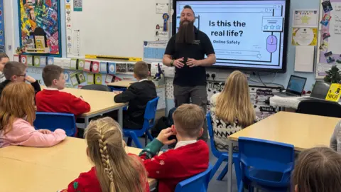 A man standing at the front of a classroom talking to children about internet safety