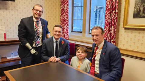 Lord Mackinlay of Richborough stands next to a table where Wes Streeting, Richard Whitehead and Tony Hudgell are all seated. The setting is a traditionally decorated room with gold and red themes on the wallpaper and curtains.