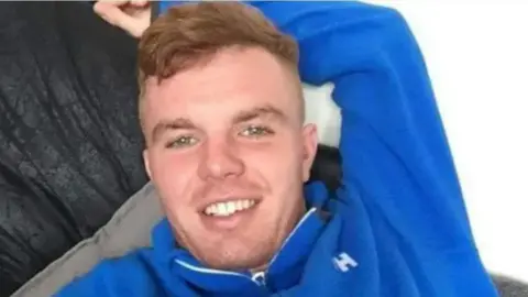 Jack O'Brien, wearing a royal blue fleece long-sleeved top, smiles at the camera as he relaxes on a sofa.