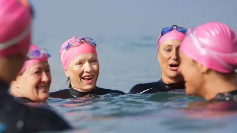 Five women are in the sea, wearing either wetsuits or rash vests - unable to tell from picture. only their heads and shoulders are visible. They wear pink swimming caps, and blue googles. They are roughly between the ages of 40 and 60.