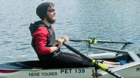 Alex Innes rowing in a boat. He is wearing glasses, a black hat, a black and red coat, and has both of his hands on oars. He has brown hair and a beard and is smiling. The boat is white and has "Nene Tourer" written on the side.