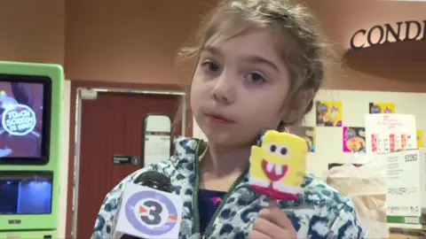 A young girl holds a Spongebob ice cream as she looks into the camera with a microphone held to her face.