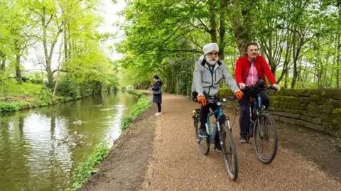 Two men on bikes cycle along a canal towpath, as a woman stands feeding ducks on the canal. There are lots of trees and a stone wall at the side. One man wears a grey coat and a turban, the other is wearing a pink and red top. 