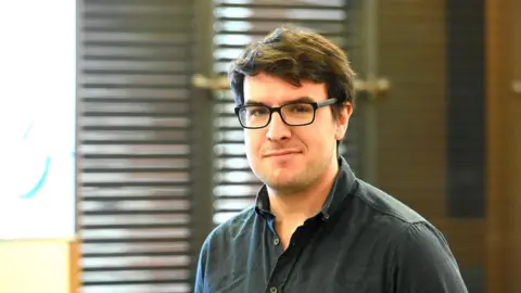 National Autistic Society A portrait of a man with dark hair and glasses wearing a dark shirt. He is standing in front of venetian blinds. 