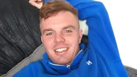 Jack O'Brien wearing a royal blue fleece long-sleeved top smiling with his arm behind his back lying on a sofa.