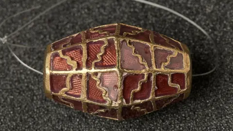 A red bead with gold details is pictured resting on a surface. A plastic loop is fitting through the bead.