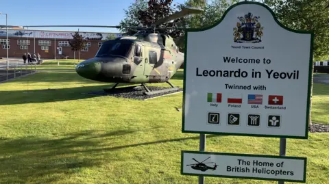 A military helicopter alongside a sign reading 'Welcome to Leonardo in Yeovil' in the foreground, while the factory building is in the background