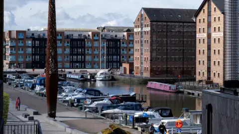 A wide shot of Gloucester Docks showing boats moored in the foreground and warehouse buildings in the background