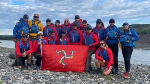 The Expedition Limitless group of 17 people wearing red coats and blue life jackets, hats and sunglasses. Half of the group is standing, the other half is kneeling in front holding a red Manx flag featuring the triskelion in white, grey and yellow. They are on a pebbled riverbank with hills in the background and a blue sky with white clouds above.