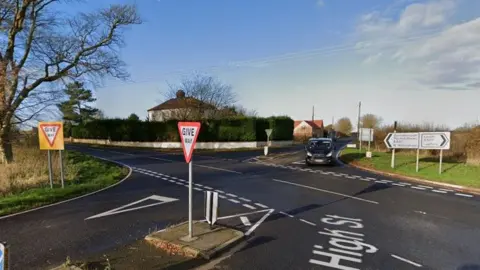 The junction of the A631 and B1225 between Horncastle and Caistor, the road has been newly tarmacked and has road markings and several give way signs. It is a rural road with fields and trees surrounding it, as well as a few houses. 