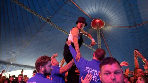 Slowthai standing above an audience under a large blue tent. He is wearing black dungarees and a black bucket hat.