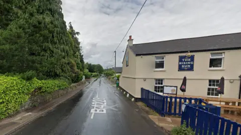 Google GV of the B4228 in Woodcroft. The road is close to a wall and trees to one side. The other side of the road is The Rising Sun Pub. It is a cream building with a blue fence.  