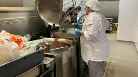 Celia using big stainless steel heavy duty kitchen machinery wearing her white cooking overalls and grey trousers