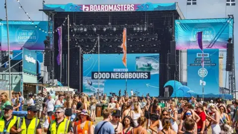 The main stage at Boardmasters in the sunshine, the stage is pale blue and reads 'Boardmasters' at the top in purple and white writing and  'up next Good Neighbours' in the centre of it. The audience are talking as they wait for the next performance.