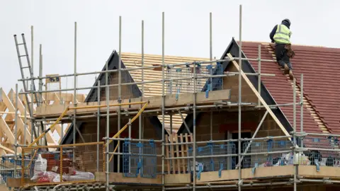 Lone builder on roof of house being built surrounded by scaffolding and wooden frames