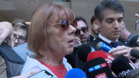 Gisèle Pélicot speaking to reporter with crowd of microphones in front of her