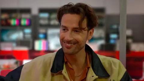 A photo of an actor called Tim Renouf. He is in a BBC studio. He is looking off towards the right and smiling. He has brown curly hair. He is wearing a black and cream jacket and an orange shirt.