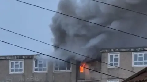 Smoke can be seen billowing into the sky from the top floor of the building. Orange flames can be seen in the staircase.