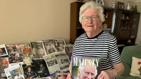 A woman holding a magazine featuring King Charles III on the cover. On the table behind her are various newspaper clippings featuring the Royal Family