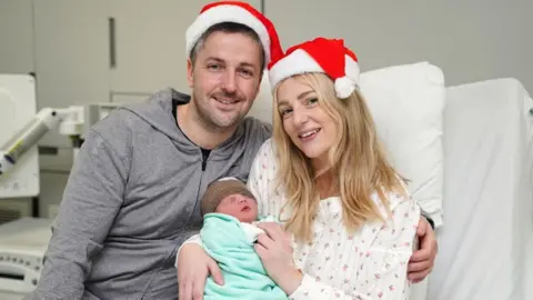 A smiling couple wearing Santa hats hold their newborn baby, wrapped in a blue blanket, while sitting in a hospital bed