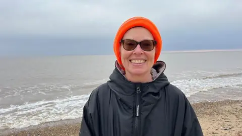 Andrew Turner/BBC Pam Spychal, dressed in a high visibility orange knitted hat, and a black dry robe. She wears glasses and is pictured on the beach at Great Yarmouth.