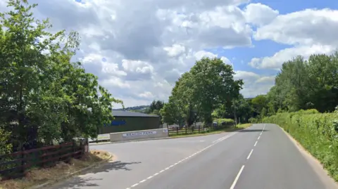 Google Google image of the entrance to Beacon Park near Dartington. Green hedges and trees line either side of the road outside of the entrance. A large white sign states Beacon Park. 