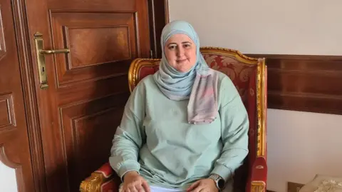 Hania is sitting in a chair, smiling at the camera. She is wearing a pastel green top and a pastel blue headscarf. 