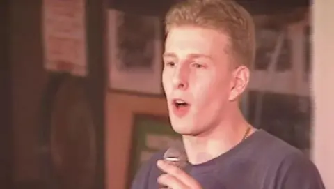 Patrick Kielty wears a navy t-shirt and is holding a mic